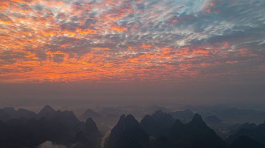 Sis kaplı karst dağlarının üzerinde canlı turuncu ve kırmızı renkleri ile gökyüzünü aydınlatan çarpıcı bir gün doğumunun nefes kesici hava manzarası. Durgun manzara, vadiler ve nehirlerle benekli.,