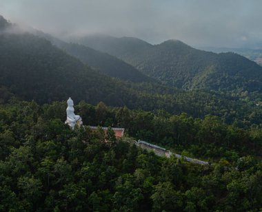 Bereketli yeşilliklerle çevrili ve uzak köyleri olan huzurlu bir vadiye bakan ormanlık bir tepeye tünemiş büyük beyaz Buda heykelinin huzurlu bir hava görüntüsü. Huzurlu ortam ruhani ve doğal uyum yayar..  