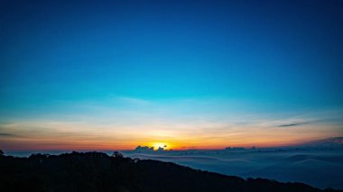 Güneş sık bir ormanın arkasında batıyor, dalgalı bulutlar ve sislerin arasından altın bir parıltı saçıyor. Bu büyüleyici sahne sıcak ve serin tonlarla harmanlanarak dramatik ve sakin bir doğal manzara yaratır..