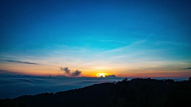 Güneş sık bir ormanın arkasında batıyor, dalgalı bulutlar ve sislerin arasından altın bir parıltı saçıyor. Bu büyüleyici sahne sıcak ve serin tonlarla harmanlanarak dramatik ve sakin bir doğal manzara yaratır..
