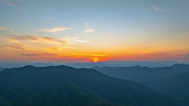 Güneş sık bir ormanın arkasında batıyor, dalgalı bulutlar ve sislerin arasından altın bir parıltı saçıyor. Bu büyüleyici sahne sıcak ve serin tonlarla harmanlanarak dramatik ve sakin bir doğal manzara yaratır..