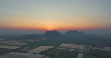 Uçsuz bucaksız pirinç tarlasının uçsuz bucaksız manzarası. Güneş, sabah gökyüzünde turuncu bir parıltı oluşturan büyük bir dağın arkasında parlıyor. Pirinç tarlasında güneş ışınlarının yansıması.