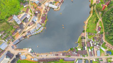 Göl kenarındaki bir köyün havadan görünüşü, kırsal mimari, yemyeşil ve sakin sulara dağılmış teknelerin karışımı sergileniyor. Doğal güzellik ve insan yerleşiminin ahenkli bir karışımı.