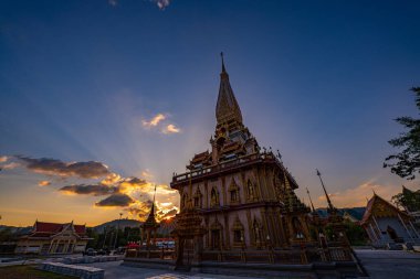 A stunning view of a traditional Thai temple Wat Chalong with intricate golden architecture, captured during sunset with dramatic clouds and warm sunlight creating a serene and spiritual atmosphere clipart