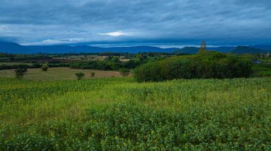 Parlak günbatımı gökyüzünün altında uzun, renkli bir dağ sırasının ve bir ayçiçeği tarlasının güzel manzarası. Bu görüntü, kırsal kesimin doğal güzelliğini, sükunetini ve çekiciliğini yansıtıyor..