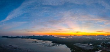 Gün batımında Phuket City 'de bir sahil şehrinin çarpıcı panoramik görüntüsü, gökyüzünde turuncu ve mavi renkli canlı renkler sergiliyor, dağlar ve kentsel ışıklar doğal ortamın uyumlu bir karışımını oluşturuyor.