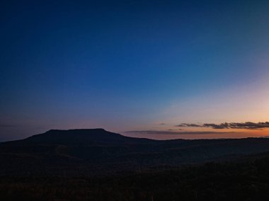 Şafakta, parlak turuncu ve pembe renkleri bulutları mavi gökyüzüne doğru aydınlatan sersemletici bir dağ manzarası.