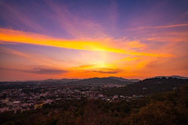 Gün batımında yemyeşil dağların arasına yerleşmiş bir kasabanın manzarası. Tepenin yamacına tünemiş bir tapınak yapısı ve canlı turuncu bir gökyüzü ılık, sakin bir atmosfer yaratıyor..