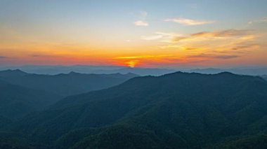 Gökyüzünde turuncu, pembe ve mavi harmanlanmış ılık renkleri ile katmanlanmış dağ siluetlerinin üzerinde soluk kesici bir gün batımı manzarası..