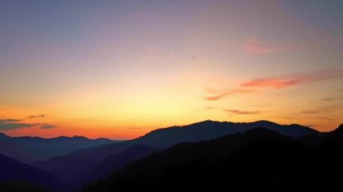 Gökyüzünde turuncu, pembe ve mavi harmanlanmış ılık renkleri ile katmanlanmış dağ siluetlerinin üzerinde soluk kesici bir gün batımı manzarası..