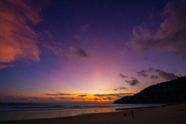 Tropikal bir sahilde nefes kesici bir gün batımı, gökyüzünde altın renkleri yansıtıyor ve sakin okyanus sularını yansıtıyor. Sahilde yürüyen insanların siluetleri huzur ve huzur katıyor. 