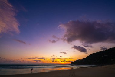 Tropikal bir sahilde nefes kesici bir gün batımı, gökyüzünde altın renkleri yansıtıyor ve sakin okyanus sularını yansıtıyor. Sahilde yürüyen insanların siluetleri huzur ve huzur katıyor. 