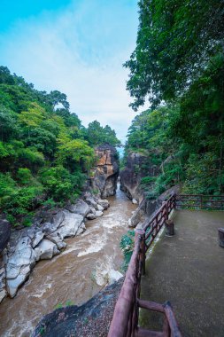 Yükselen kaya oluşumları ve uçurumları birbirine bağlayan dar bir köprüyle, yemyeşil ormanlarla çevrili bir nehir vadisinin pitoresk görüntüsü. Doğal güzellik ve canlı yeşillik. Öpücük uçurumu