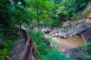 Yükselen kaya oluşumları ve uçurumları birbirine bağlayan dar bir köprüyle, yemyeşil ormanlarla çevrili bir nehir vadisinin pitoresk görüntüsü. Doğal güzellik ve canlı yeşillik. Öpücük uçurumu