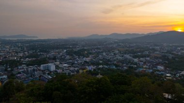 Gün batımında yemyeşil dağların arasına yerleşmiş bir kasabanın manzarası. Tepenin yamacına tünemiş bir tapınak yapısı ve canlı turuncu bir gökyüzü ılık, sakin bir atmosfer yaratıyor..