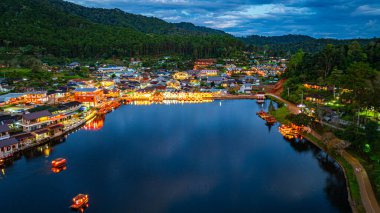 Baan Rak Thai Köyü, gün batımında Çin tarzı mimaride göl kenarındaki ev ve restoranların renkli ışıklarla yansıması.. 