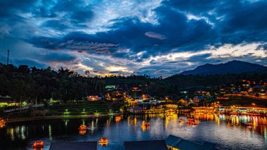 Baan Rak Thai Köyü, gün batımında Çin tarzı mimaride göl kenarındaki ev ve restoranların renkli ışıklarla yansıması..