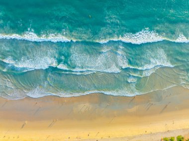 Çarpıcı bir hava manzarası turkuaz okyanus dalgalarını yakalıyor altın kumlu bir plaja doğru yavaşça yuvarlanıyor ve insanlar kıyı boyunca dağılıyorlar. Canlı renkler ve doğal dokular huzur verir. 