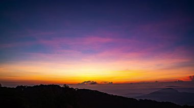 Güneş sık bir ormanın arkasında batıyor, dalgalı bulutlar ve sislerin arasından altın bir parıltı saçıyor. Bu büyüleyici sahne sıcak ve serin tonlarla harmanlanarak dramatik ve sakin bir doğal manzara yaratır..