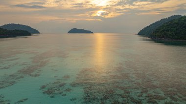 Surin Adaları üzerinde çarpıcı bir günbatımının havadan görüntüsü. Sakin okyanus sularına yansıyan altın renklerle. Sahil şeridindeki mercan resifi derinlik katıyor. Şnorkel için cennet