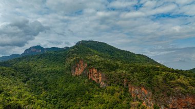 Bereketli yeşil tepelerin ve vadilerin dolambaçlı dağ yollarıyla kesiştiği hava panoramik manzarası. Manzarada engebeli uçurumlar, yoğun ormanlar ve parçalı bulutlu bir gökyüzünün altında uzak bir ufuk bulunur.. 