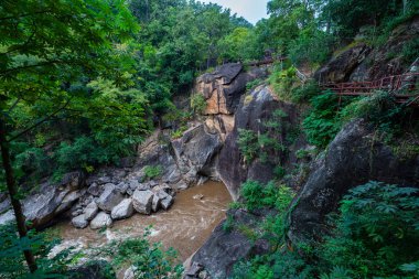 Yükselen kaya oluşumları ve uçurumları birbirine bağlayan dar bir köprüyle, yemyeşil ormanlarla çevrili bir nehir vadisinin pitoresk görüntüsü. Doğal güzellik ve canlı yeşillik. Öpücük uçurumu