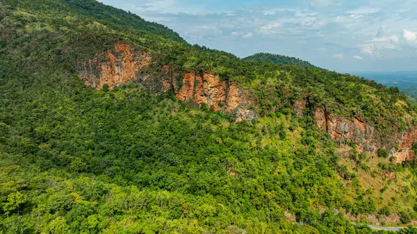 Bereketli yeşil tepelerin ve vadilerin dolambaçlı dağ yollarıyla kesiştiği hava panoramik manzarası. Manzarada engebeli uçurumlar, yoğun ormanlar ve parçalı bulutlu bir gökyüzünün altında uzak bir ufuk bulunur.. 