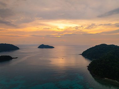 Gün batımında sakin okyanus sularıyla çevrili tropikal adaların nefes kesici bir görüntüsü. Gökyüzünün altın renkleri suyu yansıtıyor, sakin ve resimsi bir deniz manzarası yaratıyor. Surin Adası