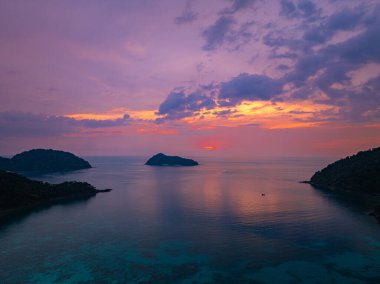 Gün batımında sakin okyanus sularıyla çevrili tropikal adaların nefes kesici bir görüntüsü. Gökyüzünün altın renkleri suyu yansıtıyor, sakin ve resimsi bir deniz manzarası yaratıyor. Surin Adası
