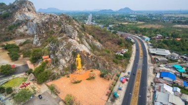 Görkemli ayakta duran altın Buda heykeli, doğal yeşillik ve uzak sıradağlarla çevrili kayalık bir kayalık yamaçta oyulmuştur. Kutsal bölgenin ruhani atmosferinin havadan görünüşü..