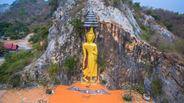 Görkemli ayakta duran altın Buda heykeli, doğal yeşillik ve uzak sıradağlarla çevrili kayalık bir kayalık yamaçta oyulmuştur. Kutsal bölgenin ruhani atmosferinin havadan görünüşü..