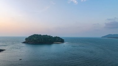 Gün batımında sakin okyanus sularına yansıyan altın rengi ile huzurlu bir sahil adasının çarpıcı hava manzarası. Ağaçların silueti ve kayalık oluşumlar huzur sahnesine derinlik katıyor..