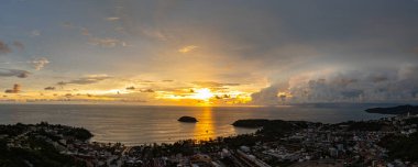 Nefes kesen bir günbatımı, kıyı kasabasının üzerinde turuncu ve mavi renkli canlı renkler çiziyor. Sahneyi çevreleyen yemyeşil tepeler var. Uzak bir tepe yapısı huzur dolu bir manzara ekler..  