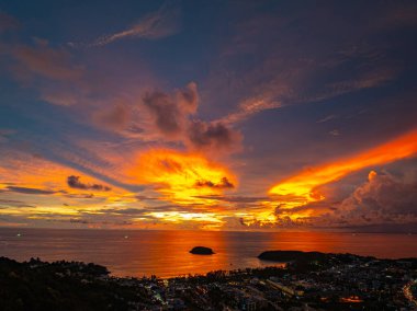 Nefes kesen bir günbatımı, kıyı kasabasının üzerinde turuncu ve mavi renkli canlı renkler çiziyor. Sahneyi çevreleyen yemyeşil tepeler var. Uzak bir tepe yapısı huzur dolu bir manzara ekler..  