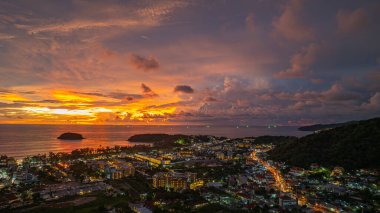 Nefes kesen bir günbatımı, kıyı kasabasının üzerinde turuncu ve mavi renkli canlı renkler çiziyor. Sahneyi çevreleyen yemyeşil tepeler var. Uzak bir tepe yapısı huzur dolu bir manzara ekler..  