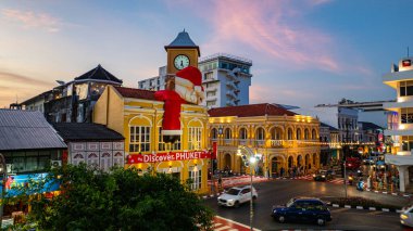 Phuket, Tayland - 30 Aralık 2024: Büyük Noel Baba ve Bayan Noel Baba süslemeleriyle süslenmiş tarihi sarı saat kulesinin olduğu canlı bir sahne dramatik bir akşam gökyüzü. Mutlu yıllar 2025