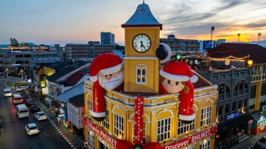 Phuket, Tayland - 30 Aralık 2024: Büyük Noel Baba ve Bayan Noel Baba süslemeleriyle süslenmiş tarihi sarı saat kulesinin olduğu canlı bir sahne dramatik bir akşam gökyüzü. Mutlu yıllar 2025