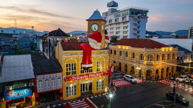 Phuket, Tayland - 30 Aralık 2024: Büyük Noel Baba ve Bayan Noel Baba süslemeleriyle süslenmiş tarihi sarı saat kulesinin olduğu canlı bir sahne dramatik bir akşam gökyüzü. Mutlu yıllar 2025