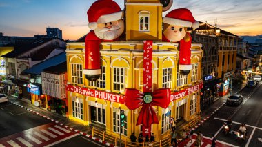Phuket, Tayland - 30 Aralık 2024: Büyük Noel Baba ve Bayan Noel Baba süslemeleriyle süslenmiş tarihi sarı saat kulesinin olduğu canlı bir sahne dramatik bir akşam gökyüzü. Mutlu yıllar 2025