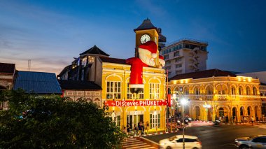 Phuket, Tayland - 30 Aralık 2024: Büyük Noel Baba ve Bayan Noel Baba süslemeleriyle süslenmiş tarihi sarı saat kulesinin olduğu canlı bir sahne dramatik bir akşam gökyüzü. Mutlu yıllar 2025