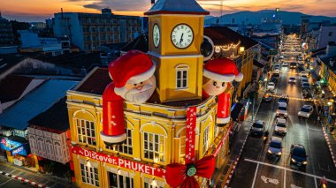 Phuket, Tayland - 30 Aralık 2024: Büyük Noel Baba ve Bayan Noel Baba süslemeleriyle süslenmiş tarihi sarı saat kulesinin olduğu canlı bir sahne dramatik bir akşam gökyüzü. Mutlu yıllar 2025