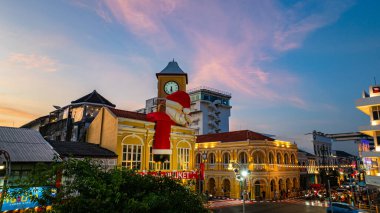 Phuket, Tayland - 30 Aralık 2024: Büyük Noel Baba ve Bayan Noel Baba süslemeleriyle süslenmiş tarihi sarı saat kulesinin olduğu canlı bir sahne dramatik bir akşam gökyüzü. Mutlu yıllar 2025