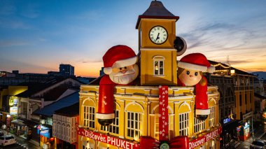 Phuket, Tayland - 30 Aralık 2024: Büyük Noel Baba ve Bayan Noel Baba süslemeleriyle süslenmiş tarihi sarı saat kulesinin olduğu canlı bir sahne dramatik bir akşam gökyüzü. Mutlu yıllar 2025