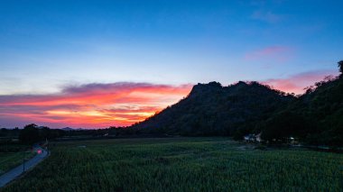 Kırmızı, turuncu ve pembe harmanlanmış alevli tonlarıyla canlı bir günbatımı gökyüzüne karşı duran dramatik dağ tepelerinin panoramik bir görüntüsü. Dağların etrafındaki ayçiçeği tarlaları.