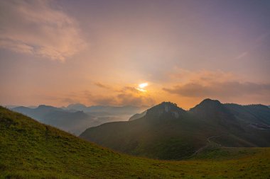 Sakin günbatımının havadan görünüşü, yuvarlanan tepeler ve uzak dağ tepeleri üzerinde altın bir parıltı oluşturarak, sisli ve yumuşak ışık katmanlarından oluşan barışçıl ve atmosferik bir manzara yaratır. dağ arkaplanı.