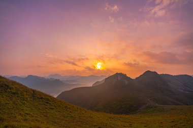 Sakin günbatımının havadan görünüşü, yuvarlanan tepeler ve uzak dağ tepeleri üzerinde altın bir parıltı oluşturarak, sisli ve yumuşak ışık katmanlarından oluşan barışçıl ve atmosferik bir manzara yaratır. dağ arkaplanı.