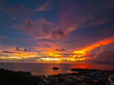 Nefes kesen bir günbatımı, kıyı kasabasının üzerinde turuncu ve mavi renkli canlı renkler çiziyor. Sahneyi çevreleyen yemyeşil tepeler var. Uzak bir tepe yapısı huzur dolu bir manzara ekler..  