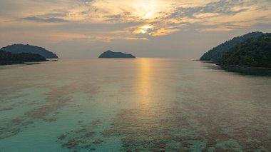 Surin Adaları üzerinde çarpıcı bir günbatımının havadan görüntüsü. Sakin okyanus sularına yansıyan altın renklerle. Sahil şeridindeki mercan resifi derinlik katıyor. Şnorkel için cennet