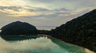 Surin Adaları üzerinde çarpıcı bir günbatımının havadan görüntüsü. Sakin okyanus sularına yansıyan altın renklerle. Sahil şeridindeki mercan resifi derinlik katar..