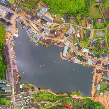 Göl kenarındaki bir köyün havadan görünüşü, kırsal mimari, yemyeşil ve sakin sulara dağılmış teknelerin karışımı sergileniyor. Doğal güzellik ve insan yerleşiminin ahenkli bir karışımı.